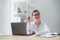 Happy young woman psychologist sits table with laptop and holds pen in her raised hand.