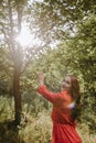 Happy young woman in red dress and with red long curly hair reaching out her hands to the sun in the forest Royalty Free Stock Photo