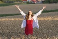 Happy young woman in red sundress and white shirt stands in the middle of wheat field with hands up. Sense of freedom