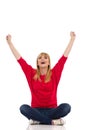 Happy Young Woman In Red Sweater Is Sitting On A Floor With Arms Raised. Front View