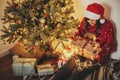 happy young woman in santa hat opening gift box at golden beautiful christmas tree with lights and presents in festive room. Royalty Free Stock Photo