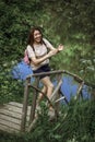 A happy young woman with short hair and a pink backpack stands on a small wooden bridge near a river in the woods, smiling and Royalty Free Stock Photo