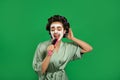 Happy young woman in silk bathrobe with hair curlers and facial mask singing into hairbrush on green background Royalty Free Stock Photo