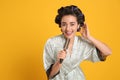 Happy young woman in silk bathrobe with hair curlers singing into hairbrush on orange background, space for text Royalty Free Stock Photo