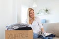 Happy young woman sit on couch stuck clothes in donation box at home, caring biracial female volunteer put apparel in carton Royalty Free Stock Photo