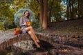 Happy young woman sitting on bringe by pond in autumn park under transparent umbrella during rain. Fall season walk Royalty Free Stock Photo