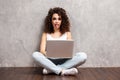 Happy young woman sitting on the floor with crossed legs and using laptop on gray background. Royalty Free Stock Photo