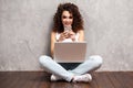 Happy young woman sitting on the floor with crossed legs and using laptop on gray background. Royalty Free Stock Photo