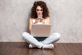 Happy young woman sitting on the floor with crossed legs and using laptop on gray background. Royalty Free Stock Photo