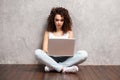 Happy young woman sitting on the floor with crossed legs and using laptop on gray background. Royalty Free Stock Photo