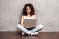 Happy young woman sitting on the floor with crossed legs and using laptop on gray background. Royalty Free Stock Photo