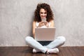 Happy young woman sitting on the floor with crossed legs and using laptop on gray background. Royalty Free Stock Photo