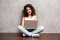 Happy young woman sitting on the floor with crossed legs and using laptop on gray background. Royalty Free Stock Photo