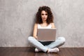 Happy young woman sitting on the floor with crossed legs and using laptop on gray background. Royalty Free Stock Photo