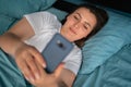Happy young woman with smartphone texting message or reading social media in bed at home. Technology, communication and Royalty Free Stock Photo