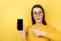 Happy young woman in spectacles, points at smart phone device, shows blank screen for your promotional content, wears sweater