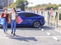 Happy young woman standing on city parking near electric car, charging automobile battery from small city station, holding shoppi