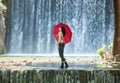 Happy young woman standing in a creek with a red umbrella in front of an impressive water fall