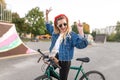 Happy young woman stands with a bicycle on the background of a skate park and rejoices with raised hands. Joyful hipster girl Royalty Free Stock Photo
