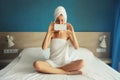 Happy young woman taking selfie with phone drying her wet hair after bath with wrapped towel on her head sitting on bed in the Royalty Free Stock Photo