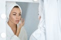 A happy young woman in a towel in front of a mirror applies cream to her face, a concept of skin care at home