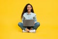 Happy young woman using laptop pc for online work or studies, sitting cross legged on yellow studio background Royalty Free Stock Photo