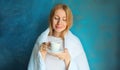 Happy young woman waking up after sleeping wrapped in white soft comfortable blanket holds cup of coffee in morning on blue