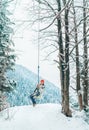 A happy young woman in warm clothes and Red Cap swinging on a forest tree swing with picturesque snowy mountain view. Wintertime Royalty Free Stock Photo