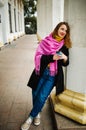 Happy young woman is wearing a pink scarf, She is holding a coffee to go. Portrait of smiling girl in autumn city. Royalty Free Stock Photo