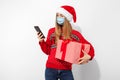 Happy young woman, wearing a red Santa Claus hat and a medical protective mask on her face, uses a mobile phone and holds a gift Royalty Free Stock Photo