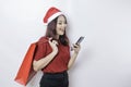 A happy young woman wearing Santa Claus` hat is smiling while holding Christmas presents and her smartphone Royalty Free Stock Photo