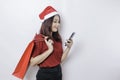 A happy young woman wearing Santa Claus` hat is smiling while holding Christmas presents and her smartphone Royalty Free Stock Photo