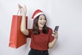 A happy young woman wearing Santa Claus` hat is smiling while holding Christmas presents and her smartphone Royalty Free Stock Photo