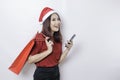 A happy young woman wearing Santa Claus` hat is smiling while holding Christmas presents and her smartphone Royalty Free Stock Photo