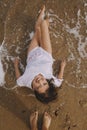 Happy young woman in wet white shirt lying on beach in splashing waves. Top view. Stylish tanned girl relaxing on seashore and