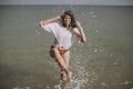 Happy young woman in wet white shirt splashing water drops with leg in sea. Stylish girl having fun in waves on beach, relaxing on Royalty Free Stock Photo