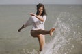 Happy young woman in wet white shirt splashing water drops with leg in sea. Stylish girl having fun in waves on beach, relaxing on Royalty Free Stock Photo