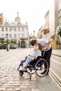 Happy young woman in wheelchair and her husband kissing her forehead, walking outdoors in old city Royalty Free Stock Photo