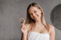 Happy young woman wrapped in white towel holding hair brush and smiling at camera, posing in bathroom interior