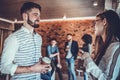 Happy young woman and young man holding cups of coffee and discussing something while standing together near the wall in office