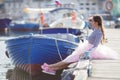 A girl in a striped sailor`s t-shirt and pink skirt sits alone on a wooden pier near the sea and boats Royalty Free Stock Photo