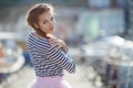 A girl in a striped sailor`s t-shirt and pink skirt sits alone on a wooden pier near the sea and boats