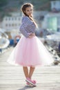 A girl in a striped sailor`s t-shirt and pink skirt sits alone on a wooden pier near the sea and boats Royalty Free Stock Photo