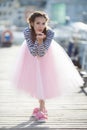 A girl in a striped sailor`s t-shirt and pink skirt sits alone on a wooden pier near the sea and boats Royalty Free Stock Photo