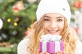 Happy youth blonde girl holds a well-packed present with a Christmas tree behind her
