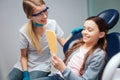 Hapy girl sit in dental chair in room. She look in mirror and smile. Female dentist sit beside her. She smile too.