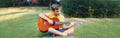 Hard of hearing preteen boy playing guitar outdoor. Child with hearing aids in ears playing music and singing song in a park.