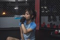 A hardworking female MMA trainee takes a seat on the ring cage floor as she hydrates herself with an electrolyte drink.