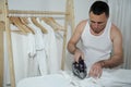 Hardworking man dressed in a white t-shirt ironing shirt on ironing board taking care of clothes, doing household chores