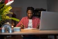 Hardworking young African guy student writing notes while watching webinar on laptop at home